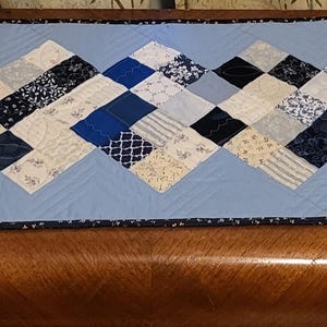 May include: A quilted table runner with a light blue background and a geometric pattern of blue, white, and black squares and rectangles. The runner has a dark blue and white patterned border.