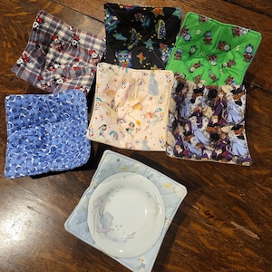 May include: A collection of colorful bowl cozies with various patterns, including Mickey Mouse, princesses, and floral designs. One cozy holds a white plate with a floral pattern. The cozies are square-shaped and quilted, providing a heat-resistant surface.