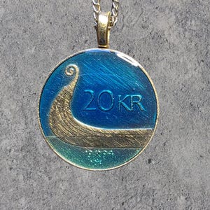 May include: A gold-toned pendant necklace featuring a round, blue and gold coin design. The coin displays a stylized boat and the text "20KR" and "1994". The pendant hangs from a gold-colored chain.