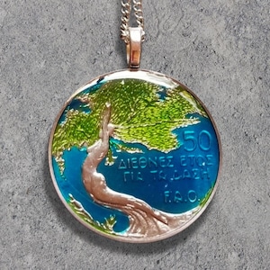 May include: Silver-toned pendant necklace featuring a round, enameled coin design. The coin depicts a tree with green leaves and a blue background, with Greek text and the number 50. The pendant hangs from a delicate chain.
