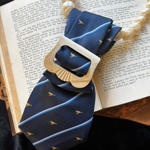 May include: Navy blue tie with a repeating pattern of yellow kangaroos and white and light blue stripes. The tie is paired with a gold-colored rectangular buckle and rests on an open book.