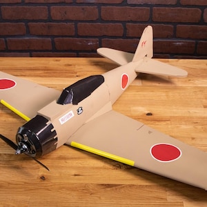 Puede incluir: Un modelo de avión de cartón beige con marcas rojas y blancas. El avión tiene una hélice negra y una franja amarilla en el ala.