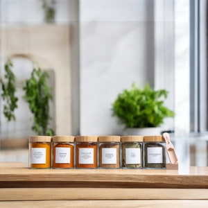 May include: Seven glass spice jars with wooden lids and labels. The labels are white with black text and list the spices: Curry Powder, Peppercorns, Kashmiri Chili Powder, Garam Masala, Dill, Fenugreek. A wooden spoon is on the right side of the image.