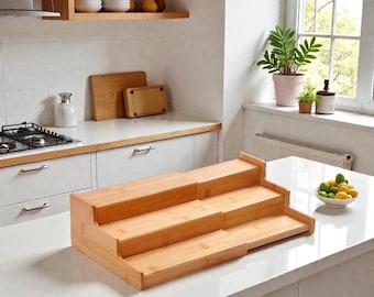 May include: A tiered bamboo spice rack with three levels. The rack is made of light-colored bamboo and is designed to sit on a countertop. The kitchen setting includes a white countertop, a window, and other kitchen items.