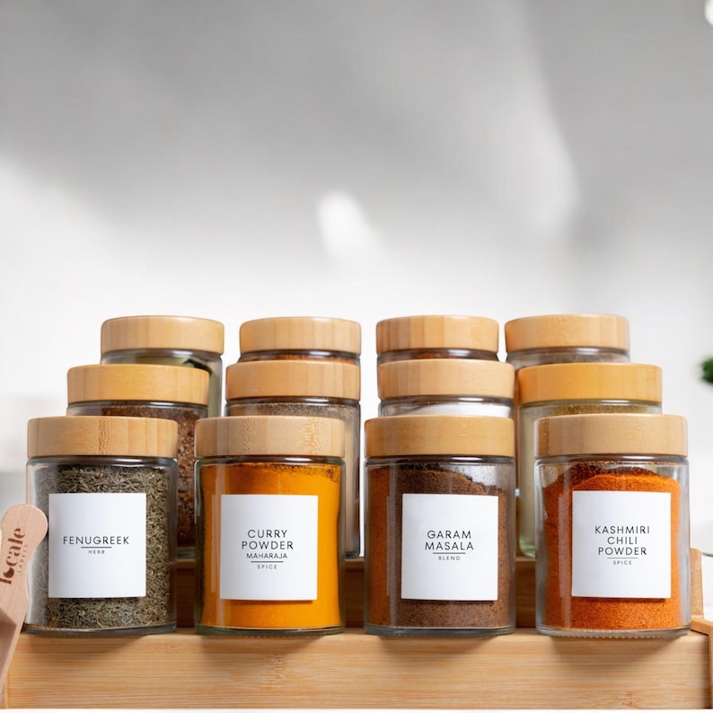 May include: A set of ten glass spice jars with wooden lids. The jars are filled with various spices and labeled with the names of the spices, including Fenugreek, Curry Powder, Garam Masala, and Kashmiri Chili Powder.