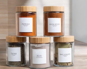 May include: Six glass spice jars with bamboo lids. The jars are filled with various spices and herbs, including paprika, suya spice, thyme, achi, and bay leaves. Each jar has a white label with the name of the spice or herb printed in black.