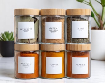 May include: Six glass spice jars with bamboo lids are stacked on a white surface. Each jar contains a different spice and has a white label with the spice name. Spices include bay leaves, garam masala, thyme, achiote powder, curry, and paprika.
