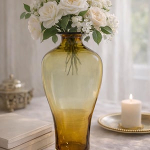 May include: A tall, amber-colored glass vase holds a bouquet of white roses and smaller white flowers with green leaves. A lit candle sits on a gold-colored plate. The vase is on a white tablecloth.