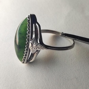 Op de afbeelding: Een zilveren ring met een groene edelsteen gezet in een decoratieve zilveren setting. De ring heeft een bloemmotief op de band.
