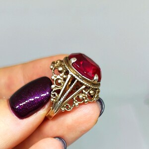 May include: A vintage gold-toned ring featuring a large, faceted, rectangular red gemstone. The ring has an ornate filigree design around the gemstone and on the band. The ring is held up by a hand with dark purple nail polish.