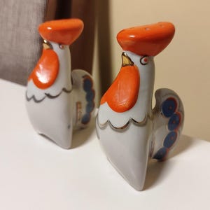 May include: Two ceramic rooster salt and pepper shakers. The roosters are white with orange combs and wattles, blue and red tail feathers, and gold accents.