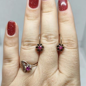 May include: A gold-toned ring and earrings set. The ring features a leaf design with a central red gemstone. The earrings have a flower design with a red gemstone center. The jewelry is set against a neutral background.
