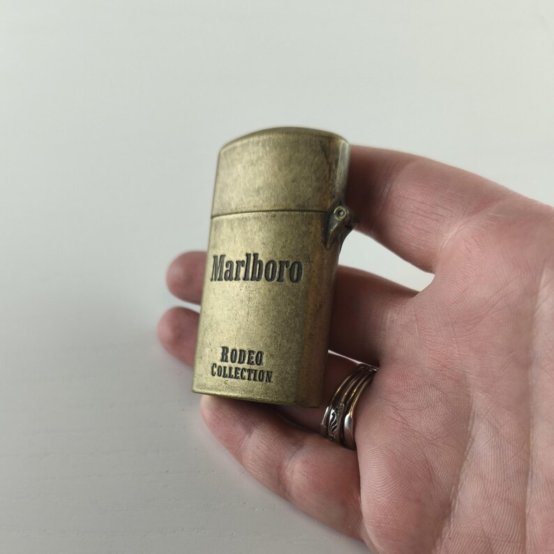 May include: A vintage brass-colored lighter with the word "Marlboro" printed on the front, along with "RODEO COLLECTION." The lighter is held in a hand, showcasing its aged appearance.