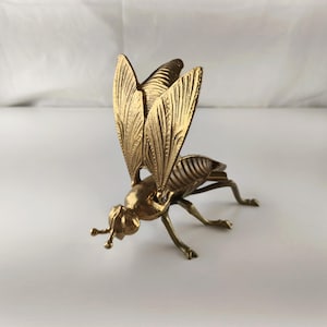 May include: A detailed brass fly figurine with outstretched wings. The insect sculpture has intricate wing and body detailing, and is posed with its legs extended. The fly is a golden color and appears to be made of metal.
