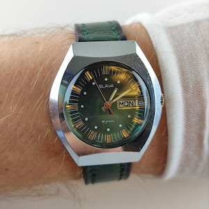 Puede incluir: Un reloj de pulsera Slava vintage con esfera verde y caja plateada. El reloj tiene una pantalla de día y fecha y una correa de cuero verde. La esfera del reloj tiene agujas e indicadores dorados. El reloj es un reloj clásico.