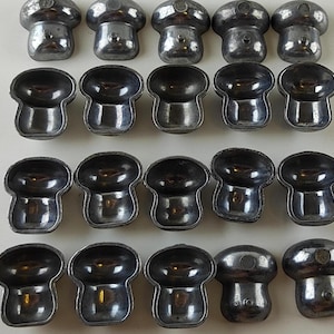 May include: A collection of twenty-one small, silver-colored metal objects. The objects have a mushroom-like shape, with a rounded top and a flared base. The tops have a small, circular design. The objects are arranged in rows and columns on a white surface.