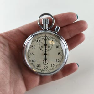 Può includere: Un cronometro vintage cromato con quadrante bianco e numeri neri. Il cronometro ha un anello in alto e un pulsante laterale. Il quadrante mostra numeri da 0 a 60, con segni dei secondi. Le parole "MADE IN USSR" sono stampate in basso.
