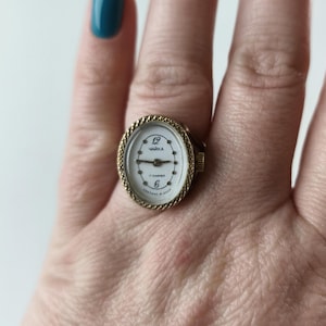 Pode incluir: Um relógio de anel dourado com mostrador oval. O mostrador do relógio é branco com algarismos e ponteiros pretos, e o texto "12 ЧАЙКА 6" é visível. O anel tem uma borda texturizada em forma de corda.