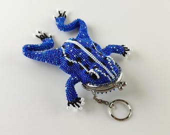 May include: A beaded frog-shaped coin purse in shades of blue, black, and white. The frog has a silver-toned clasp and a key ring attached. The beaded design creates a textured surface, and the frog's feet are detailed with white beads.
