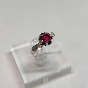 Puede incluir: Un anillo de plata con una gema grande, redonda y de color rojo intenso. La gema está engastada en un engaste oscuro y ornamentado. El anillo se exhibe sobre un soporte de acrílico transparente sobre un fondo blanco.