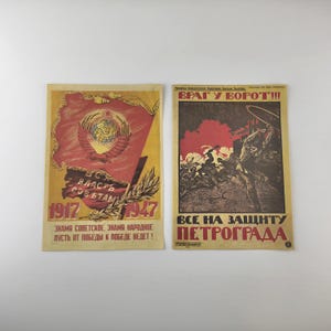 Op de afbeelding: Twee vintage Sovjet-propaganda-posters. De linkerposter toont een rode vlag met een hamer- en sikkel-embleem, de tekst "1917-1947" en Russische tekst. De rechterposter heeft een rood-bruin kleurenschema met Russische tekst en illustraties.