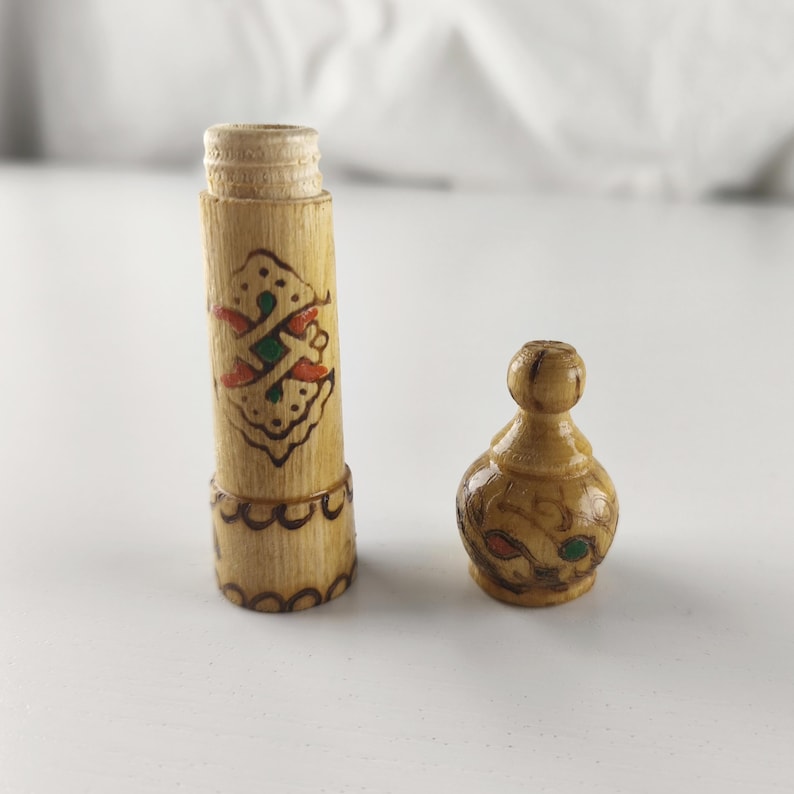 Puede incluir: Un peque&ntilde;o recipiente de madera con un dise&ntilde;o decorativo. El recipiente es cil&iacute;ndrico con una tapa de rosca. La tapa est&aacute; separada y tiene una parte superior redondeada. El recipiente est&aacute; decorado con detalles rojos, verdes y marrones.