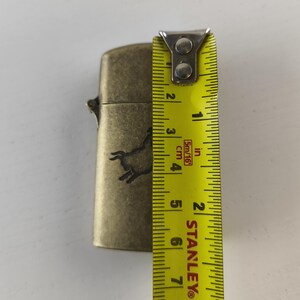 May include: A brass-colored lighter with a horse silhouette design. The lighter measures approximately 2.5 inches (6.35 cm) in height. The lighter is shown with a yellow measuring tape.