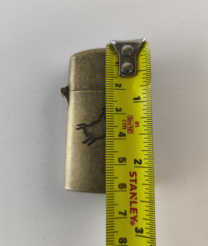 May include: A brass-colored lighter with a horse silhouette design. The lighter measures approximately 2.5 inches (6.35 cm) in height. The lighter is shown with a yellow measuring tape.
