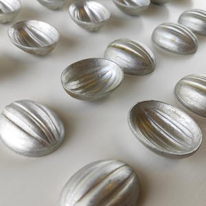 Könnte beinhalten: Eine Ansammlung silberfarbener, oval geformter Formen mit gerippten Details. Die Formen sind auf einer weißen Oberfläche angeordnet und zeigen ihren metallischen Glanz und ihr strukturiertes Design. Wahrscheinlich zum Backen oder Basteln.