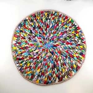 May include: A round, colorful trivet made from woven fabric strips. The trivet features a spiral design with a variety of colors, including red, yellow, blue, and green. The trivet is approximately 10 inches in diameter and has a brown border.