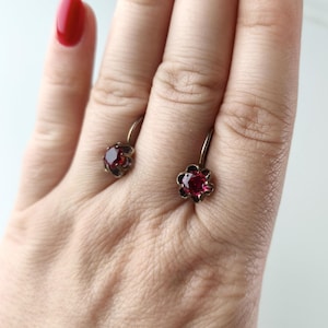 May include: A pair of earrings featuring round, deep red faceted stones set in a metallic flower-shaped frame. Each earring has a curved wire hook closure. The design highlights the contrast between the dark metal setting and the vibrant red gemstone.