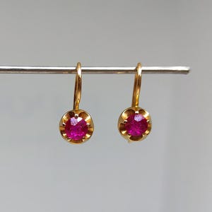 May include: A pair of gold-colored leverback earrings, each featuring a vibrant, round, ruby-red gemstone. The earrings are displayed against a neutral background, highlighting their color and design. The gemstones are set in a delicate, scalloped bezel.