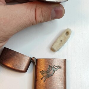 May include: A copper-coloured metal lighter with an open lid, displaying a bull-riding design. The lighter is disassembled, showing the internal components, including the wick and flint. The lighter is on a white surface.