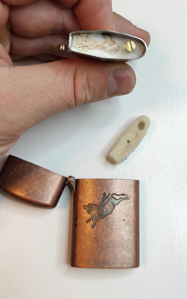 May include: A copper-coloured metal lighter with an open lid, displaying a bull-riding design. The lighter is disassembled, showing the internal components, including the wick and flint. The lighter is on a white surface.