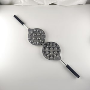 Könnte beinhalten: Ein Vintage-Nusswaffeleisen aus Gusseisen mit zwei Scharnierteilen, jeweils mit ovalen Formen. Es hat lange Metallgriffe mit schwarzen Kunststoffgriffen. Das Waffeleisen ist geöffnet und zeigt die Formen.