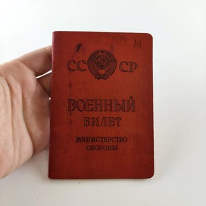 May include: A vintage red military document with Cyrillic text and the emblem of the USSR. The document, titled "Military ID," features the text "CCCP" and "Министерство Обороны" (Ministry of Defense).