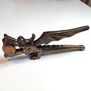 May include: A bronze dragon nutcracker with wings outstretched, holding a nut in its mouth. The nutcracker is made of metal and has a detailed design.