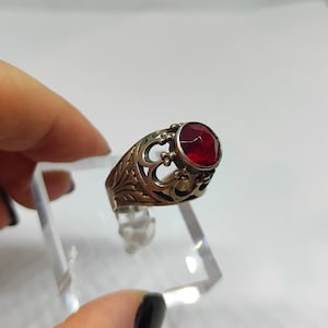 Op de afbeelding: Een antieke ring met een sierlijk, opengewerkt ontwerp. De ring heeft een grote, ronde, gefacetteerde rode edelsteen. Het metaal heeft een aangetaste, verouderde uitstraling. De ring wordt op een transparante standaard gepresenteerd.