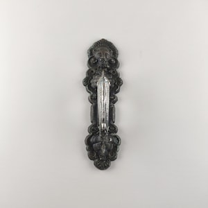 May include: Ornate, dark metal wall sconce with a central, clear glass candle holder. The sconce features a detailed design with a face at the top and floral motifs. The overall length is approximately 10 inches.