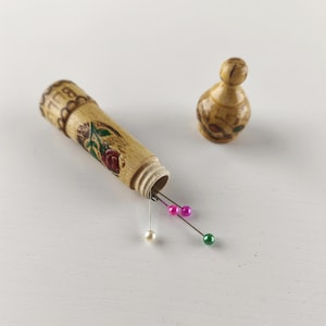 Könnte beinhalten: Ein hölzerner Nadelhalter mit floralem Design und separatem Deckel. Der Nadelhalter ist geöffnet und zeigt Nadeln mit farbigen Köpfen. Das Design umfasst eine rote Rose und grüne Blätter. Der Deckel hat eine kleine, abgerundete Form.