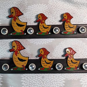 Puede incluir: Dos juegos de colgadores de pared decorativos con forma de pato. Cada juego presenta tres patos amarillos con pañuelos rojos y bases verdes, montados sobre una tabla rectangular marrón oscuro con pomos plateados.