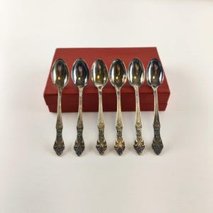 May include: Six silver spoons with ornate handles and blue accents are arranged in a row. The spoons are in front of a red box, creating a classic and elegant presentation. The spoons are likely for serving or eating.