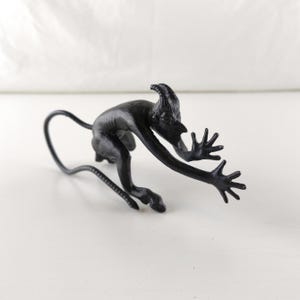 May include: A matte black figurine of a horned creature in a crouching, reaching pose. The figure has elongated limbs, splayed fingers, a pointed face, and a long, thin, curved tail extending behind it, crafted with a smooth, solid finish.