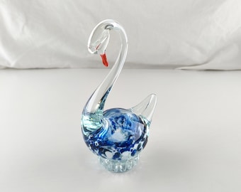 May include: A clear glass swan figurine with a long, curved neck and a red beak. The body of the swan is filled with blue and white swirls, and it sits on a clear, textured base. The swan is a decorative item.