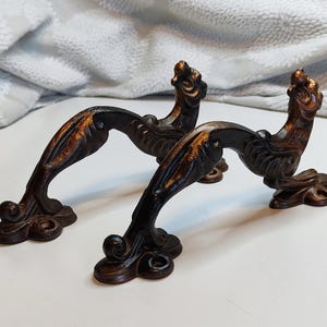 以下が含まれることがあります： ブロンズ色の金属製の装飾的なドアハンドル2つ。各ハンドルは、装飾的なベースと、上部に詳細な様式化された動物の頭が付いた曲線的なデザインが特徴です。キャビネットまたはドアに適しています。