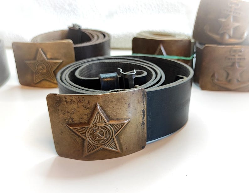 K&ouml;nnte beinhalten: Eine Sammlung von Vintage-Lederg&uuml;rteln mit Messing-Schnallen, die ein Stern- und Hammer-Symbol und einen Anker zeigen. Die G&uuml;rtel sind in verschiedenen Braunt&ouml;nen und Schwarz.