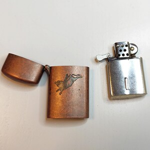 May include: Two vintage lighters. One is silver-toned with the top open. The other is copper-toned with a hinged lid open, revealing an etched design of a bull. The lighters are on a white surface.
