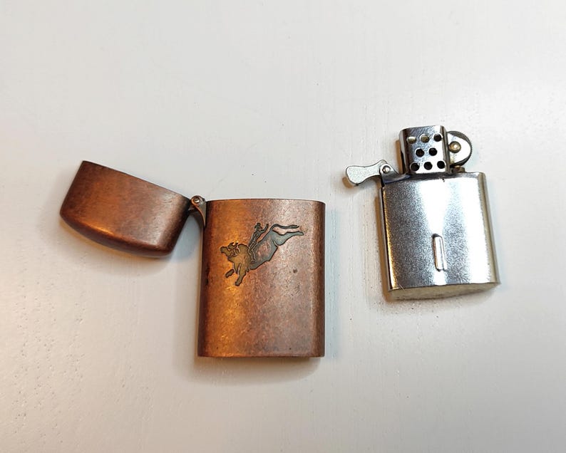 May include: Two vintage lighters. One is silver-toned with the top open. The other is copper-toned with a hinged lid open, revealing an etched design of a bull. The lighters are on a white surface.