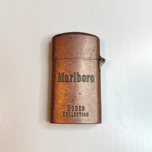 May include: A copper-toned lighter featuring the Marlboro and Rodeo Collection branding. The rectangular lighter has a hinged lid and rounded corners. The text is embossed on the front of the lighter.