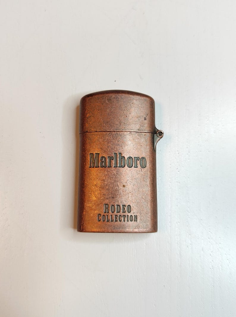 May include: A copper-toned lighter featuring the Marlboro and Rodeo Collection branding. The rectangular lighter has a hinged lid and rounded corners. The text is embossed on the front of the lighter.